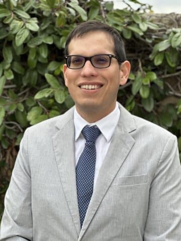 A Latino man with short brown hair and black glasses, wearing a gray suit jacket, white shirt and navy tie, smiling.