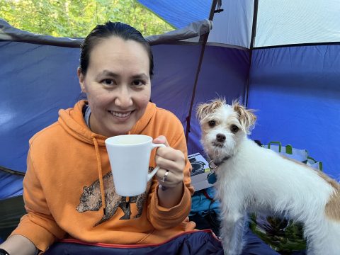Photo of Nancy Stalowski, an Asian-American woman with long black hair put in a ponytail, wearing an orange hoodie, holding a coffee mug and smiling inside a tent. Next to her is a small white and brown dog, looking at the camera and "smiling".