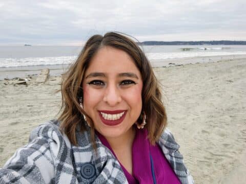 A Latina woman with short brown hair, wearing magenta lipstick and a purple shirt, with a checkered white and grey jacket smiling, in front of a beach.