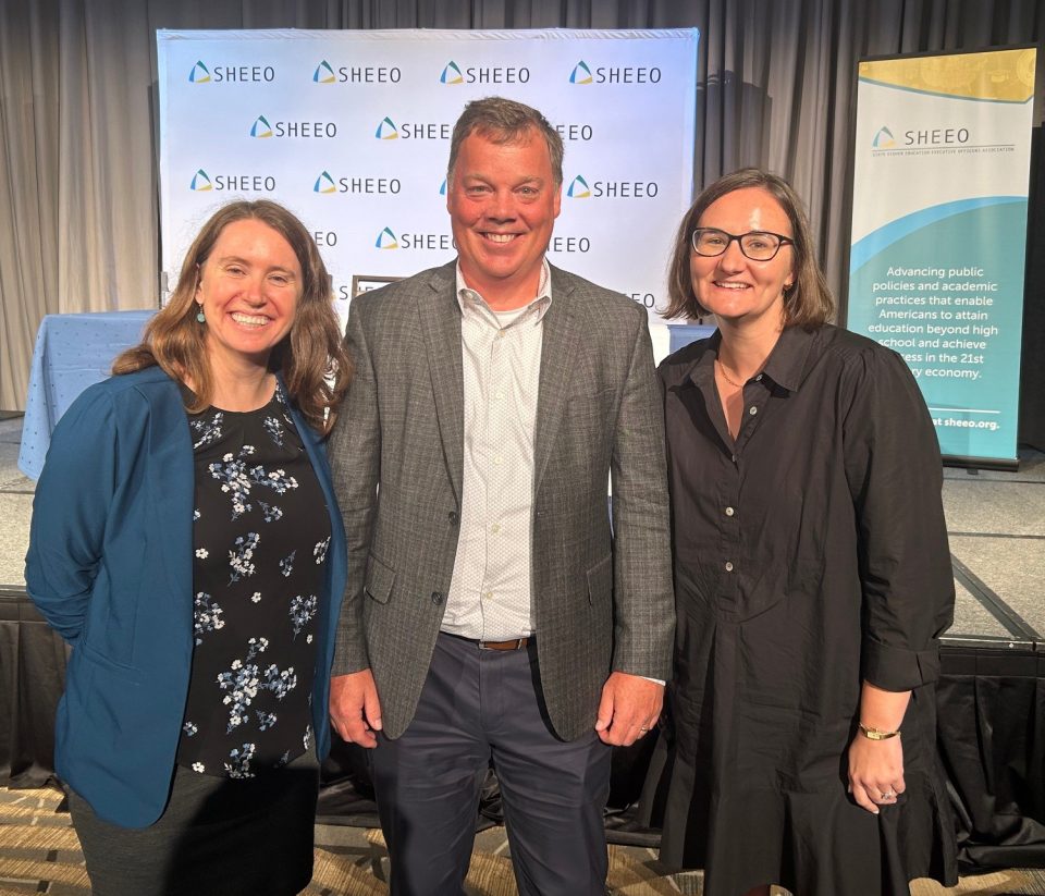 Photo of Louisa Hunkerstorm, Brian Prescott, and Sarah Pingel at SHEEO Policy Conference in front of stage with SHEEO backdrop.